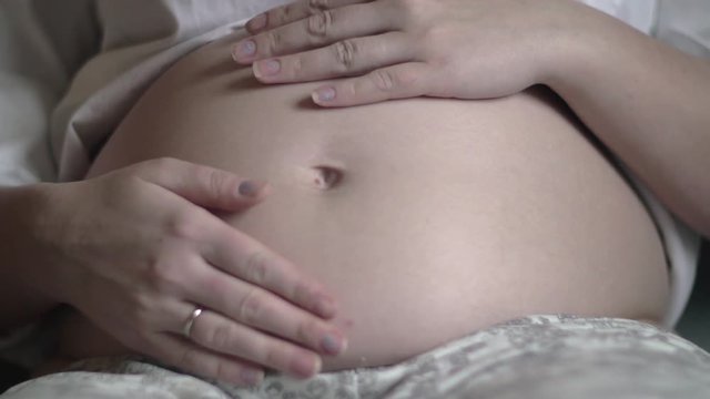 A closeup of a pregnant woman's belly. Her hands are slowly and gently touching it and making a heart shape around the belly button