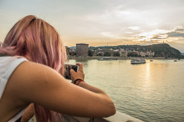 Young girl with pink hair back to camera take photo phone from Budapest waterfront urban city district in evening sunset time 