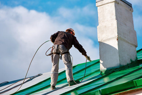 Worker of Industrial Alpinist Services painting roof in green colors with paint spray gun. Professional climber wearing uniform, helmet and using safety harness. Risky job. Extreme occupation.