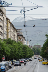 Fototapeta premium Avenue at the downtown of Sofua, Bulgaria. A lot of traffic and a lot of green trees along the road. Snowy mountain and medieval church on the background.