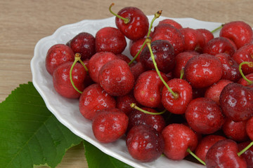 Ripe red cherries on a white plate on a wooden table. Around the green leaves.