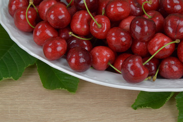 Ripe red cherries on a white plate on a wooden table. Around the green leaves.