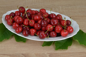 Ripe red cherries on a white plate on a wooden table. Around the green leaves.