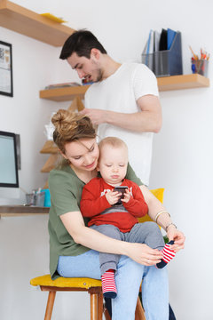 Everyday life of ordinary families. Young beautiful mother dresses socks to her small child playing smartphone against the backdrop of dad with magazine in modern interior