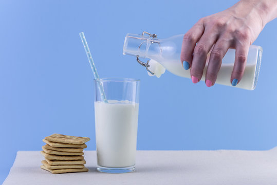 Woman's hand pours fresh milk from bottle into glass and cookies on a blue background. Concept of healthy dairy products
