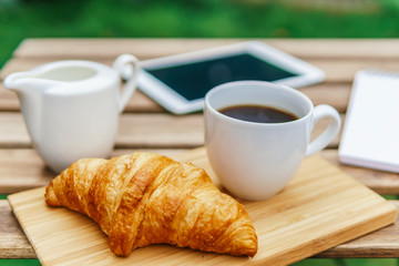 Morning Breakfast In Green Garden With French Croissant, Coffee Cup, Orange Juice, Tablet and Notes Book On Wooden Table