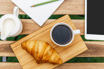 Morning Breakfast In Green Garden With French Croissant, Coffee Cup, Orange Juice, Tablet and Notes Book On Wooden Table