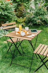 Morning Breakfast In Green Garden With French Croissant, Coffee Cup, Orange Juice, Tablet and Notes Book On Wooden Table