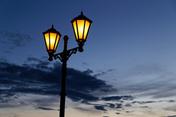 Vintage street lantern with warm yellow light and dark blue night cloudy sky on background