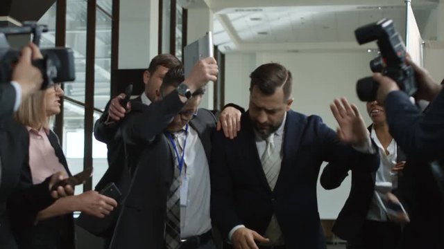Bearded formal man among immodest journalists with cameras leaving hall of conference without speech