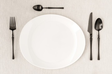 Black fork, knife, spoon and tea spoon with a white plate on a linen tablecloth.