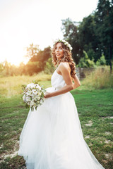Fototapeta na wymiar A cute curly woman in a white wedding dress with a wedding bouquet and wreath in her hair standing back to the camera in nature. Concept escaped bride. Forward to a happy bright future Runaway