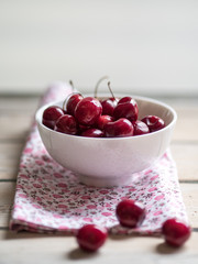White ceramic bowl of cherries on white rustic wooden table. Close up.
