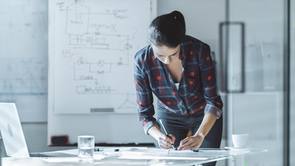 In Bright Modern Office Female Design Engineer Works on Documents, Drafts With Engineer's Scale Ruler. In the Room Whiteboard with Sketches on it, on the Walls Pinned Blueprints.