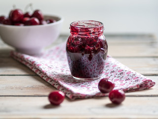 White ceramic bowl of cherries and homemade jam in jar on white rustic wooden table. Close up.
