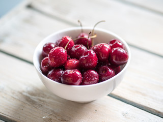 White ceramic bowl of cherries on white wooden table. Close up.