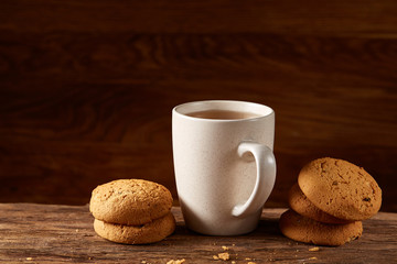 White porcelain mug of tea and sweet cookies on piece of wood over wooden background, top view, selective focus