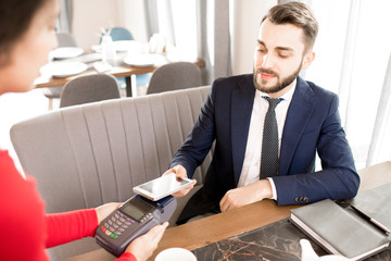 Content handsome bearded businessman with mustache using smartphone for NFC payment while waitress holding credit card reader in restaurant