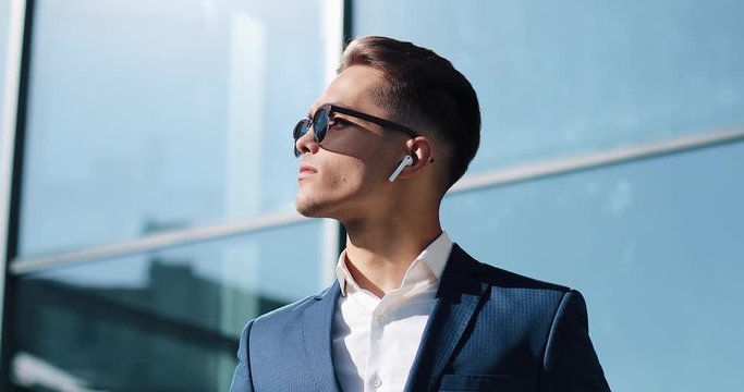 Attractive young businessman using wireless earphones standing outside. He wearing sunglasses and looks away. Communication, finance, business people, interns, CEO. Close up. Shot on Red Epic