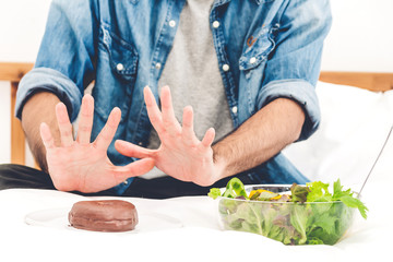 Man making choice between healthy salad and calorie bomb chocolate donut.Man rejecting the plate with unhealthy donut on bed at home.Healthy eating diet and Junk food concept