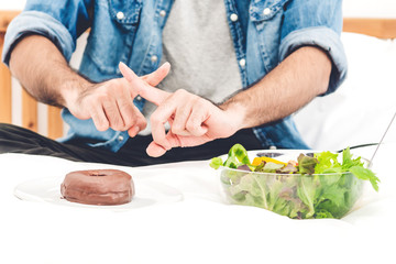 Man making choice between healthy salad and calorie bomb chocolate donut.Man rejecting the plate with unhealthy donut on bed at home.Healthy eating diet and Junk food concept