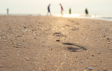 Close up view. Footprint of person walking on the sand. Shadow blur of people doing activities at the background. During the sunrise.