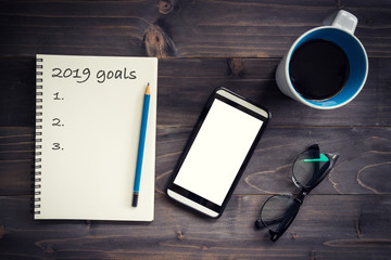 Notebook with 2019 goals massage with pencil, glasses, phone and cup of coffee on wood background.