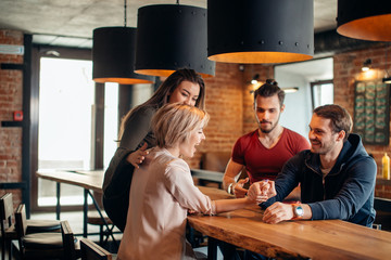 Joyful young couple in burger pub loughing and testing their strengths sitting at table doing arm wrestling with happy smiles. Their friends cheering the up