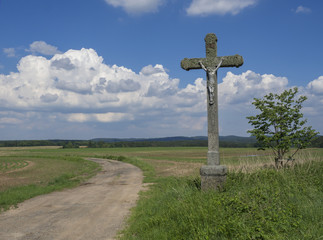 Fototapeta na wymiar idyllic rural landscape with old stone cross with Jesus Christ statue, crucifix, dirty road, green fields, hills, tree and blue sky white fluffy clouds