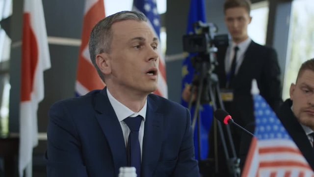 Adult male representative of America telling speech into microphone while sitting at table on international summit with other members