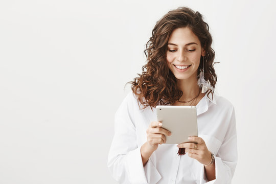 Charming cute caucasian woman in white trendy blouse browsing in net via tablet, smiling and expressing happiness, standing against gray background. Fashionable woman editing her photos in app