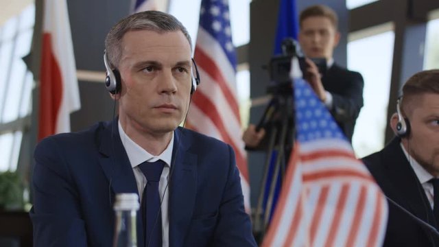 Adult man of American deputy wearing headphones and listening to speech translation while sitting at table on summit
