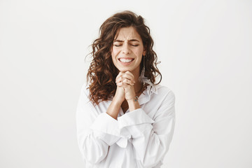 Studio shot of worried and upset caucasian woman holding hands in pray near chin, squeezing it...