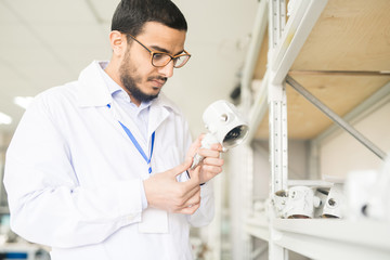 Waist-u portrait of handsome Arabian engineer wearing eyeglasses and lab coat examining quality of pressure sensor while standing at spacious warehouse of modern plant