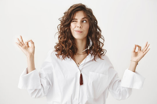 Fototapeta Studio portrait of charming positive caucasian female businesswoman trying to relax while meditating, standing with lifted hands and zen signs over gray background, peeking and looking aside playfully