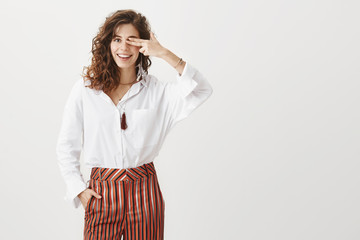 Horizontal studio shot of carefree and confident female office worker in stylish clothes, holding fingers on eye, smiling broadly, standing with hand in pocket over gray background, happy and relaxed