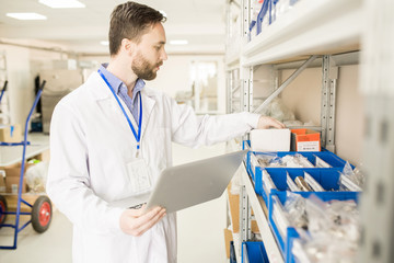 Concentrated bearded technician wearing lab coat checking existence of spare parts for  pressure sensors at spacious warehouse of modern factory and taking necessary notes on laptop.