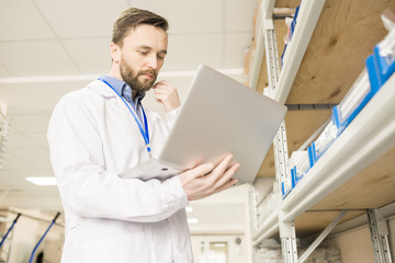 Pensive middle-aged engineer wearing lab coat using modern laptop while examining existence of fasteners in containers at spacious warehouse of modern factory
