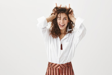 Fototapeta premium Indoor shot of feminine and flirty happy female coworker in stylish blouse and striped pants, winking and smiling while sowing tongue, holding victory or peace gesture on hair mimicking ears