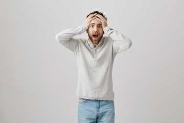 Terrified miserable bearded guy holding hands on hair while shouting and expressing anxiety and nervousness, standing over gray background. Man trembles seeing his cat walking on handrail of balcony