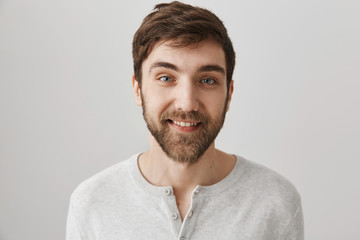 Fototapeta premium Close-up studio portrait of friendly good-looking male with beard, wearing white pullover and standing over gray background, smiling joyfully at camera. Helpful employee ready to work hard