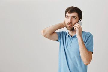 Thoughtful bothered caucasian guy with beard, talking on phone while touching neck and looking up with nervous expression over gray background. Man listens to his boss who gives extra assignments