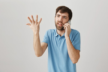 Stop boiling my brain with nonsense. Studio shot of angry european bearded guy talking on smartphone and gesturing, being irritated or fed up while standing against gray background. I lose my temper