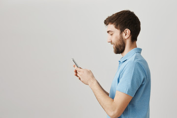 New model of phone is much better. Profile portrait of entertained happy young bearded guy holding smartphone and typing something while messaging or browsing in ner over gray background