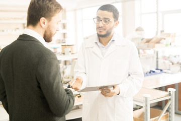 Young Arabian engineer wearing lab coat holding clipboard in hands while discussing results of accomplished work with bearded superior while standing at production department of modern factory.