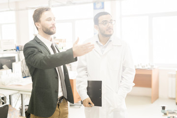 Bearded middle-aged businessman giving tour of production department of modern factory, young Arabian inspector wearing lab coat listening to him with interest