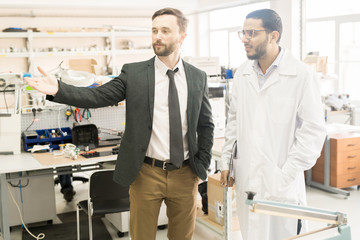 Handsome middle-aged entrepreneur in formalwear discussing working process with lab technician while standing at spacious production department of manometer factory