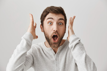 Guy screaming from lots of positive emotions hearing some unbelievable news. Portrait of overwhelmed european guy shaking hands near face and standing with opened mouth over gray wall, being surprised