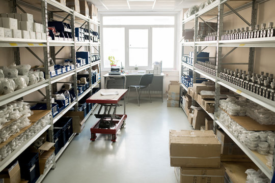 Interior of spacious warehouse of modern manometer factory: shelves with pressure sensors and spare parts, pile of cardboard boxes, wooden desk and office chair