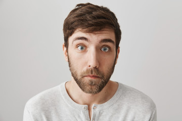 Obraz premium Close-up portrait of surprised and upset european male with beard standing with lifted eyebrows, clueless and tired expression over gray background. Guy is shocked to hear sad news